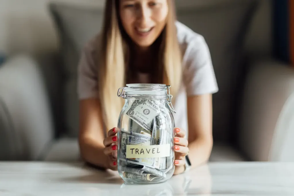 Une femme qui tient un bocal avec de l'argent à l'intérieur et l'étiquette TRAVEL sur le devant